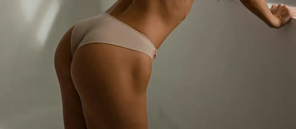 Glúteos de una mujer vista de espaldas con ropa interior clara, apoyada junto a una ventana, sin rostro